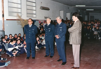 Cuatro hombres de pie, tres con uniforme azul oscuro y uno con traje beige, se presentan frente a una audiencia de niños sentados en el suelo de un gimnasio