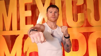 Un hombre con camiseta blanca sin mangas posa con los puños cerrados hacia el frente ante un fondo con letras de color amarillo.