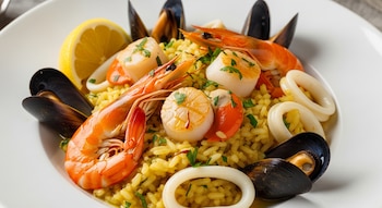 Un plato blanco contiene arroz amarillo, cuatro camarones, cuatro vieiras, anillos de calamar, mejillones, perejil picado y un gajo de limón.