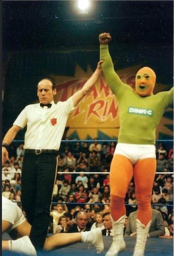 Un árbitro sostiene en alto el brazo de un luchador enmascarado con traje verde y naranja dentro de un cuadrilátero de lucha libre