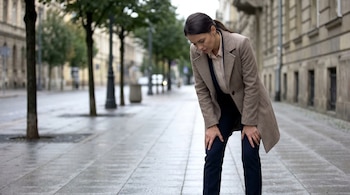 Una mujer con abrigo beige y pantalón oscuro se inclina con las manos en las rodillas sobre una vereda mojada. El fondo es una calle urbana con edificios y árboles borrosos.