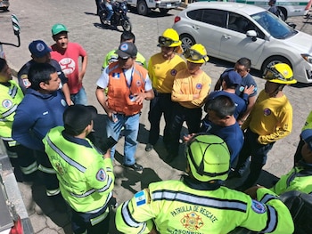 Grupo de personas con uniformes de rescate verdes y amarillos, cascos de seguridad, chaleco naranja, y banderas de Guatemala, en una reunión al aire libre
