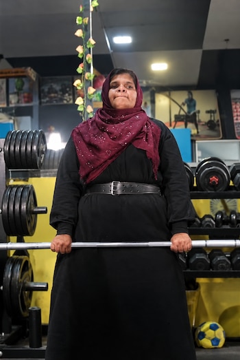 Una mujer con velo granate, ropa negra y cinturón sostiene una barra de metal frente a estantes con discos y mancuernas