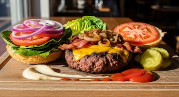 Una hamburguesa deconstruida en tabla de madera con carne, queso, tocino, cebolla caramelizada, lechuga, tomate, cebolla roja, pepinillos y salsas.