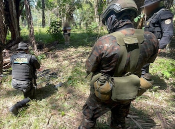 Tres agentes de seguridad uniformados en un entorno boscoso; uno arrodillado, otro de espaldas con equipo táctico, y una persona con rostro difuminado al fondo