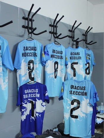 Siete camisetas de fútbol celestes y azules con los números 1, 2, 5, 8 y 9 cuelgan de ganchos en un vestuario. El texto "Gracias Selección" es visible