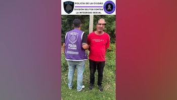 Un agente de policía con chaleco morado sujeta a un hombre de camiseta roja y rostro pixelado. Al fondo, vegetación y un letrero policial