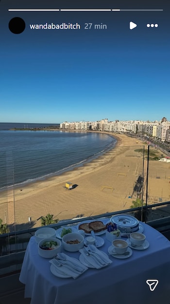 Mesa con desayuno en un balcón, con tazas de café, tostadas, frutas y cereales, frente a una playa de arena, el mar y edificios costeros bajo cielo azul claro