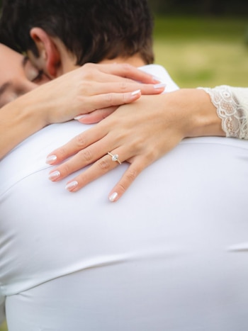 Manos de una mujer con un anillo de compromiso de diamante ovalado, abrazando la espalda de un hombre con camisa blanca. La mujer tiene uñas con esmalte nacarado