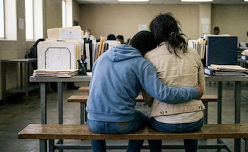 Vista trasera de dos adolescentes sentados en un banco de madera, uno abrazando al otro, en un espacio institucional con pilas de documentos en escritorios
