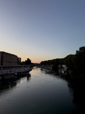 Río Tíber en Roma al anochecer, fluyendo entre edificios de la ribera izquierda y árboles de la ribera derecha. Cielo claro con colores suaves del atardecer