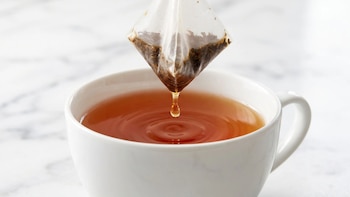 Una bolsa de té piramidal suspendida sobre una taza blanca llena de líquido ámbar con una gota que cae y genera ondas en la superficie
