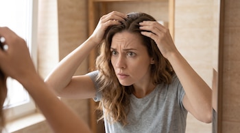 Mujer con cabello castaño rizado y camiseta gris se mira al espejo y separa mechones de cabello para observar su cuero cabelludo.