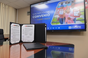 Documento abierto sobre una mesa reflectante en una oficina, con una gran pantalla de fondo que muestra el texto 'SUSCRIPCIÓN DE CONVENIO FONAFE Y ENACE' y una imagen de una familia