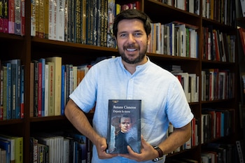 Hombre con barba y camisa clara sostiene un libro negro con el título "Renato Cisneros Dejarás la tierra" delante de estantes llenos de libros