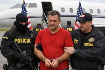 Hombre esposado con camiseta roja es escoltado por un agente policial y un Marshal de EE. UU. en un aeropuerto, con un jet privado y banderas estadounidenses.