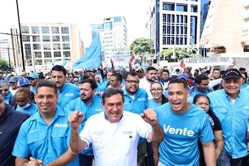 Multitud de personas camina por una calle urbana con edificios altos. Varios manifestantes visten camisetas celestes con "Vente" y sostienen banderas