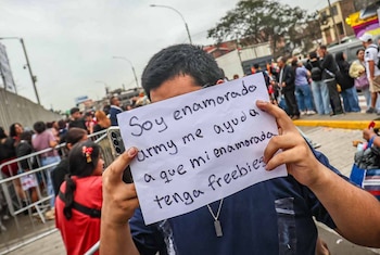 Un hombre sostiene frente a su rostro un papel blanco con un mensaje manuscrito en medio de una multitud al aire libre junto a vallas metálicas