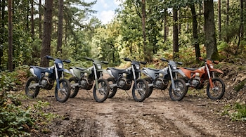 Cinco motocicletas de enduro de distintos colores alineadas en un camino de tierra rodeado de árboles.
