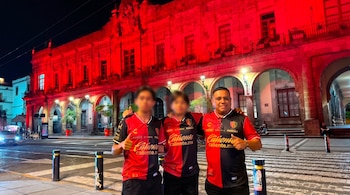 Tres personas con rostros borrosos visten camisetas rojas y negras y posan de noche frente a un edificio con arcos y ventanas iluminado con luz roja.