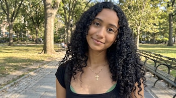 Celeste Rivas, una mujer con cabello rizado oscuro, sonríe a la cámara en un primer plano. Lleva un collar de cruz dorada. Al fondo, un parque arbolado.