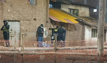 Policías y un civil observan una escena nocturna en Cusco, con edificios de adobe y techos de calamina, detrás de una valla de seguridad naranja