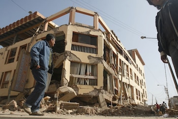 Hombre en chaqueta vaquera camina sobre escombros de construcción frente a edificio de varios pisos con daños estructurales. Un cartel dice 'TRAGAMONEDAS LA MINA'