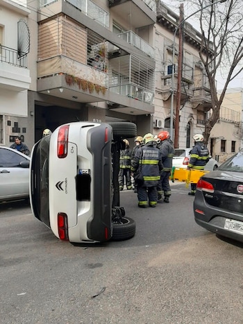 Un automóvil Citroën C4 blanco está volcado de lado sobre el asfalto. Varios bomberos con uniforme rodean el vehículo. Hay edificios y otros coches en la calle