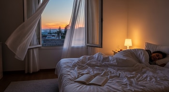 Dormitorio con cama blanca y persona durmiendo. Ventana abierta con cortinas ondeando, mostrando un atardecer. Un libro y una lámpara en mesita de noche.