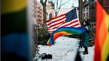 Una mujer con abrigo verde oscuro sostiene una bandera arcoíris en un paisaje nevado, con una gran bandera de Estados Unidos ondeando detrás y edificios urbanos al fondo