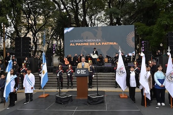 Hombre en podio, flanqueado por granaderos y niños con banderas argentinas. Detrás, orquesta y pantalla con mensaje "LA CIUDAD HOMENAJEJA AL PADRE DE LA PATRIA"