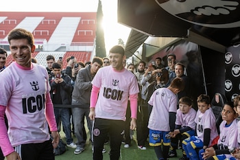 Dos futbolistas con camisetas rosadas sonríen en una cancha verde, rodeados de público y niños. Un fondo de gradas rojas y blancas, con el sol brillando