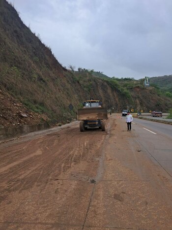 Cargador frontal amarillo en carretera cubierta de lodo; un hombre, vehículos, conos de tráfico y una ladera montañosa son visibles bajo un cielo nublado