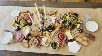 Mesa con papel kraft cubierta por una picada que incluye quesos, embutidos, uvas, frutos secos, galletas, pan, olivas, encurtidos, dulces, velas y platos.