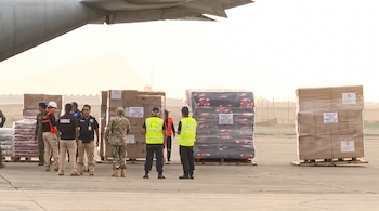 Personal militar y civil supervisa palés de cajas y bultos en pista de aeropuerto, bajo el ala de una aeronave