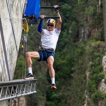 Hombre con casco y arnés colgado de barras metálicas junto a una plataforma y un muro vertical con vegetación de fondo