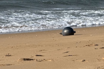 Objeto esférico de color oscuro semienterrado en la arena, con olas rompiendo en la orilla de una playa