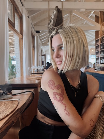 Mujer de cabello rubio con un tatuaje de dragón en el brazo derecho, sentada en la mesa de un restaurante