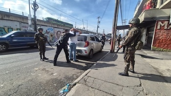 Agentes uniformados y armados junto a un automóvil compacto gris mientras registran a un hombre con las manos en la cabeza