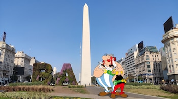 Obelisco de Buenos Aires, letras BA con vegetación, personajes de Asterix, Obelix y Dogmatix, edificios, cielo azul despejado, plantas.
