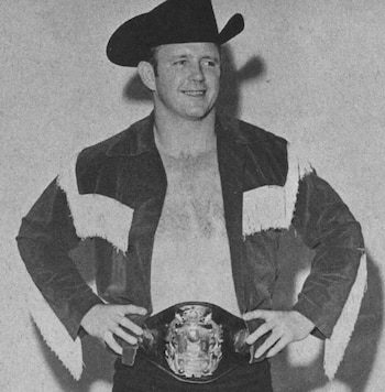 Hombre sonriente con sombrero de vaquero negro, chaqueta abierta con flecos y un cinturón de campeonato de lucha libre en la cintura