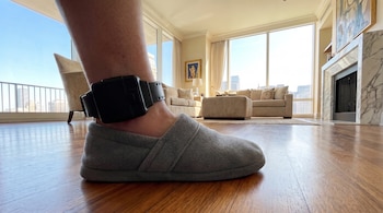 Primer plano de un tobillo con brazalete electrónico de monitoreo y una pantufla en un apartamento.