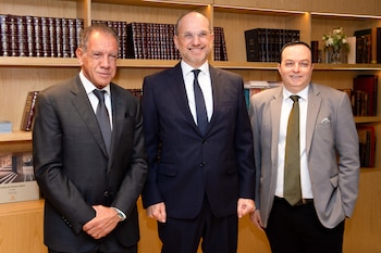 Tres hombres con traje y corbata están de pie y miran a la cámara delante de una estantería de madera con libros encuadernados