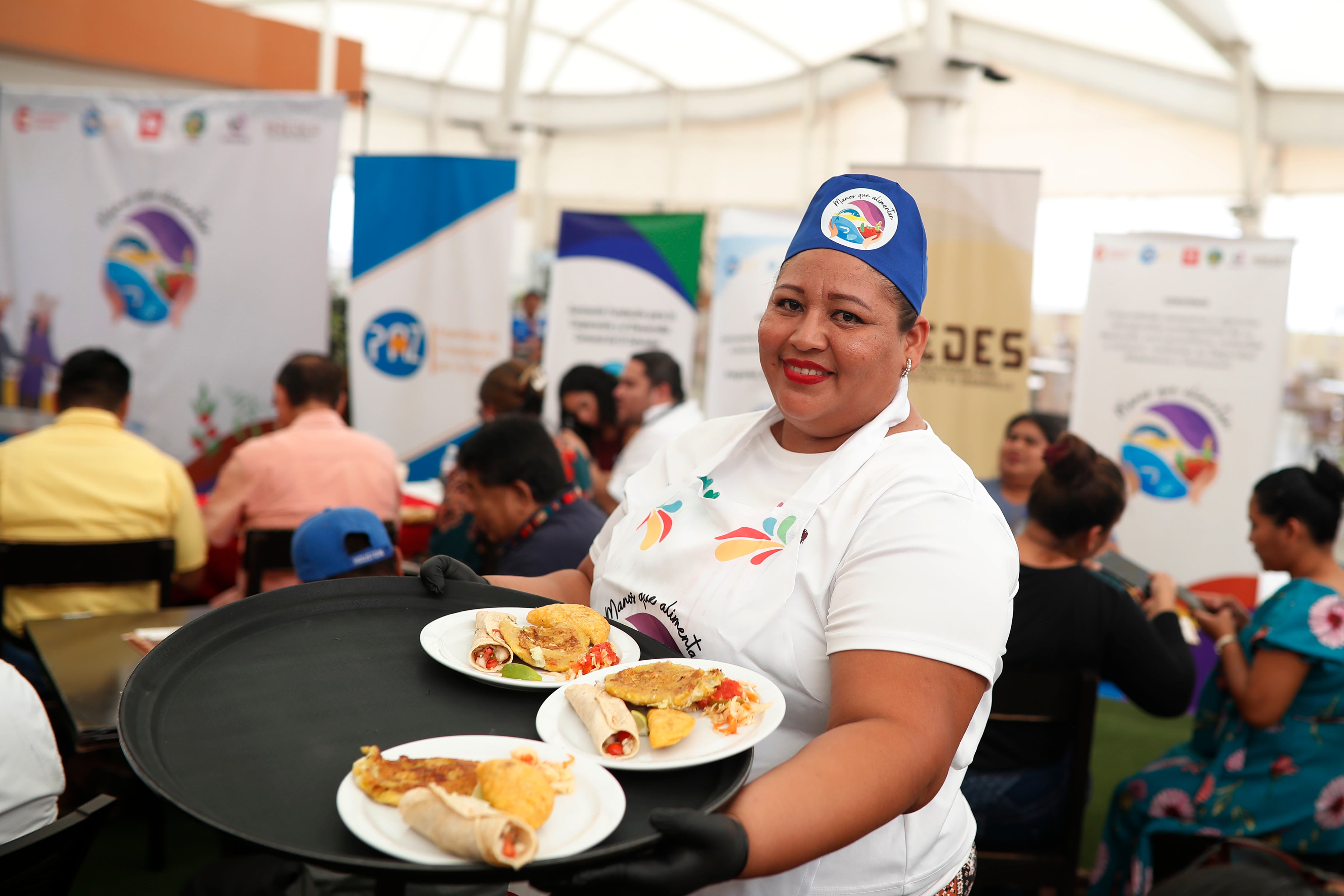 Una mujer sonríe mientras sirve platos de comida, representando la iniciativa Manos que Alimentan, financiada por Cooperación Española y liderada por Asamblea de Cooperación por la Paz, en un evento en San Salvador. (Agencia EFE)