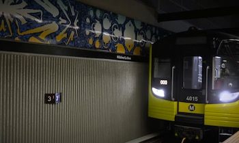 La parte frontal de un tren amarillo y negro se ve en una estación subterránea, junto a una pared con un patrón vertical y un mural artístico