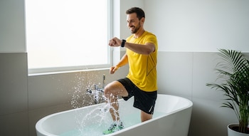Hombre sonriente con camiseta amarilla y pantalones cortos negros trotando en una bañera blanca llena de agua, con salpicaduras y un reloj inteligente en su muñeca.
