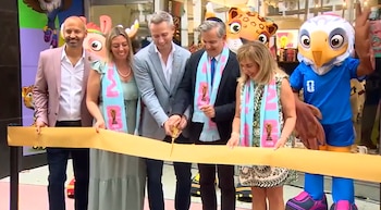 Cinco personas, incluyendo un hombre con kipá, cortan una cinta dorada en la inauguración de una tienda, con bufandas de la Copa Mundial y mascotas de fondo
