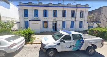 Camioneta policial blanca y automóvil estacionados en la calle frente a un edificio blanco de dos plantas con persianas azules