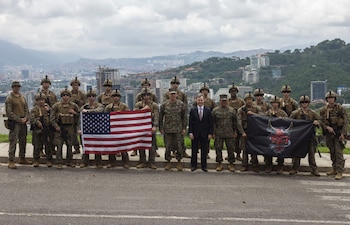 Grupo de militares estadounidenses y un civil en traje. Sostienen la bandera de EE. UU. y una bandera negra con un demonio rojo. Al fondo se ve una ciudad y montañas