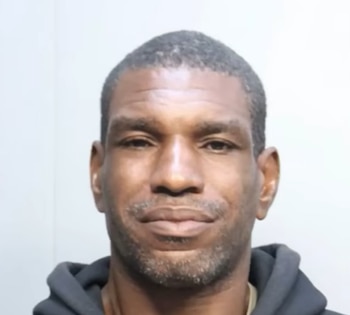 Un hombre de raza negra, de mediana edad, con barba incipiente y cabello oscuro, viste una sudadera negra sobre un fondo blanco liso